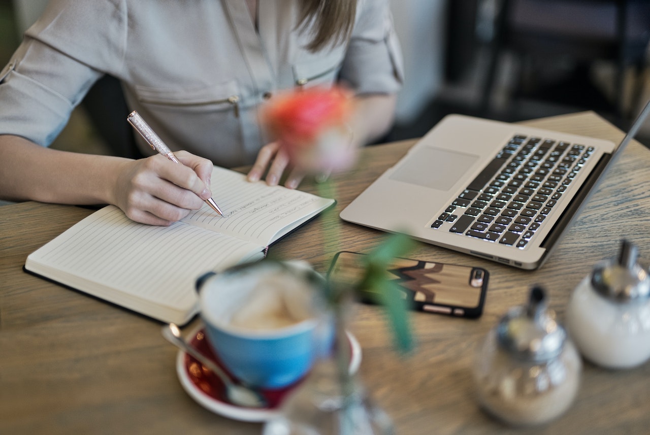 A person is writing in a notebook with a pen at a wooden table. There is an open laptop nearby, along with a smartphone, a cup of coffee, and a small vase with a flower.